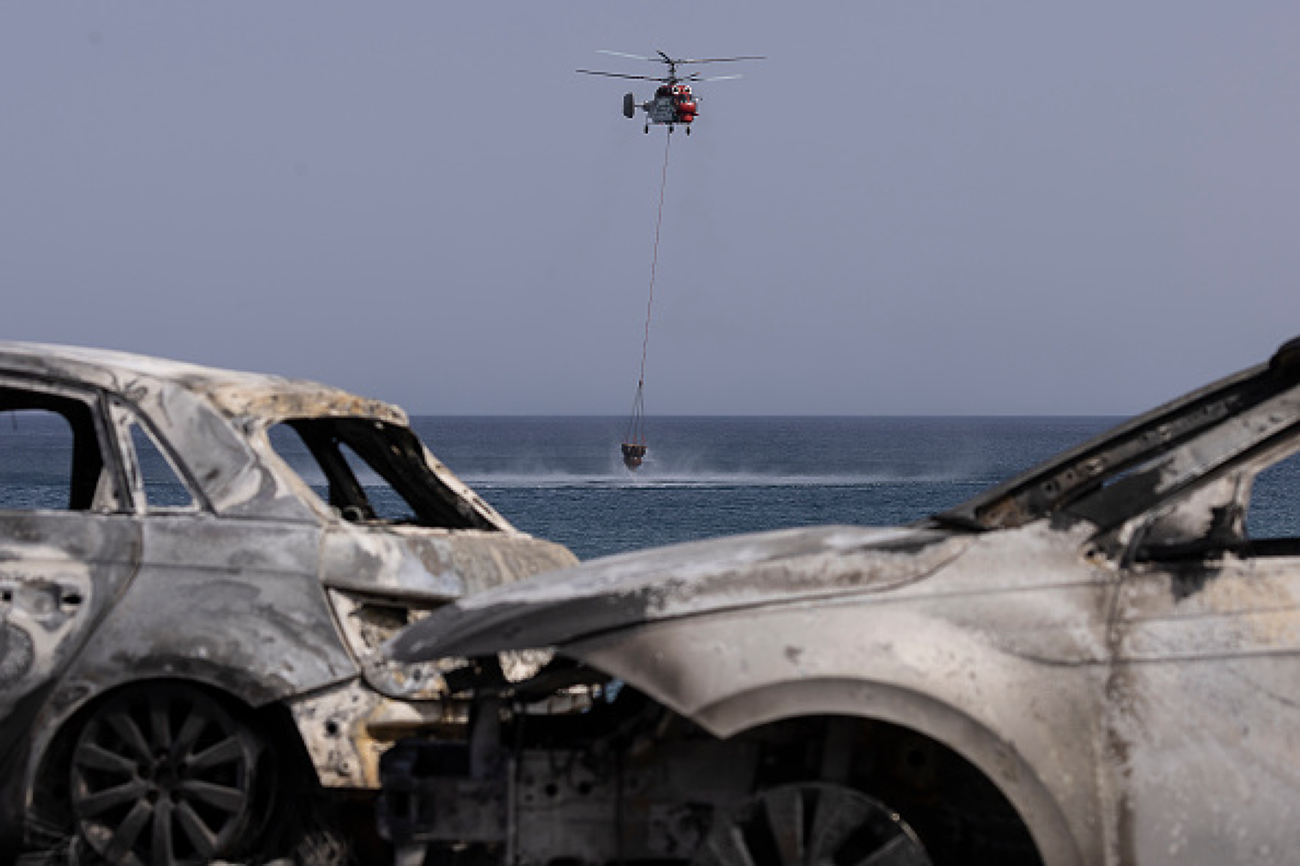 В зловеща рамка от две изгорели коли, противопожарен хеликоптер загребва вода от морето за гасене на горски пожар. Снимка: Getty Images